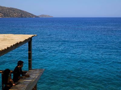Zwei Personen sitzen auf einem Holzsteg mit Blick auf das blaue Meer unter klarem Himmel.