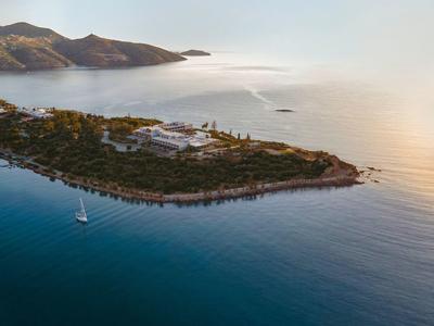 Vista aerea di una piccola isola con edifici e una barca a vela nel mare calmo al tramonto.