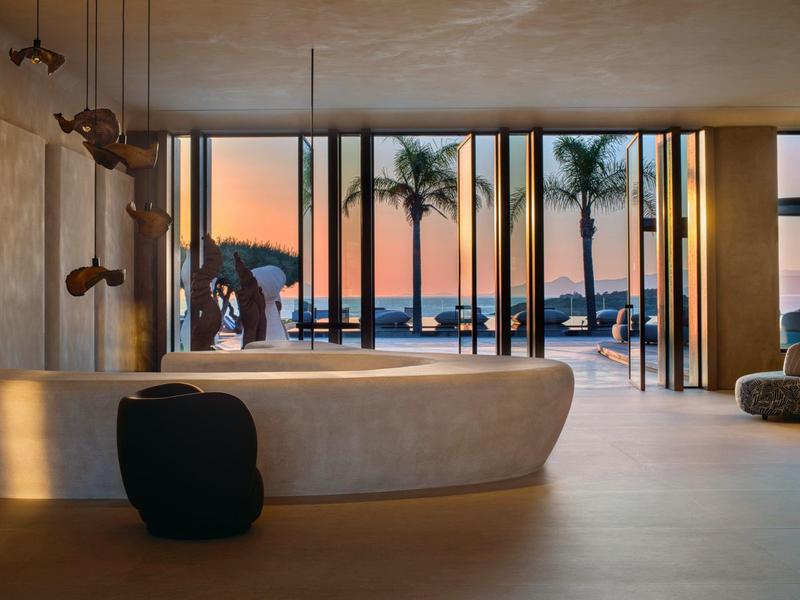 Moderne hotellobby met een organisch gevormde balie en uitzicht op palmbomen bij zonsondergang.