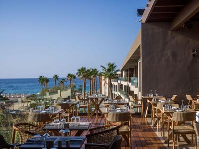 Hotel terrace with tables and chairs, overlooking palm trees and the ocean in the background.