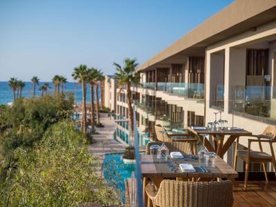 Modern hotel terrace with pool, palm trees, and sea view under clear sky.