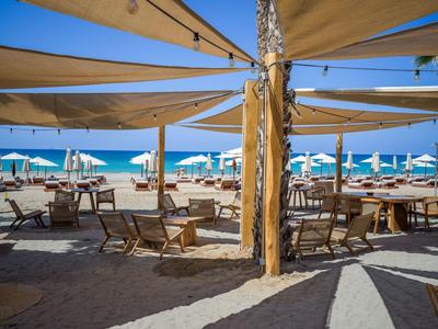 Beachside restaurant with wooden chairs, tables, and sunshades on a sunny day.