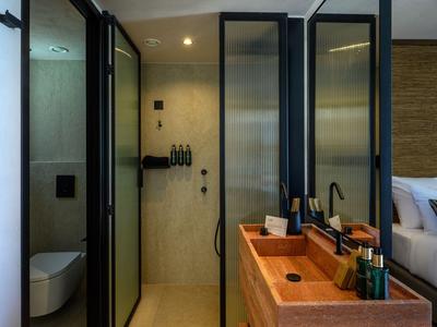 Baño moderno con ducha de vidrio, tocador de madera y habitación de invitados al fondo.
