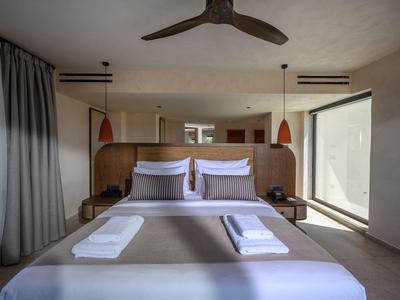Habitación de hotel moderna con cama grande, cubrecama gris, almohadas blancas y ventanas grandes.