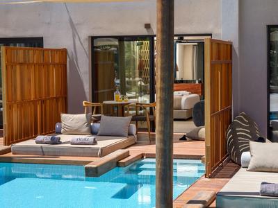 Una zona de piscina moderna con tumbonas de madera y separadores frente a grandes puertas de cristal de un hotel.
