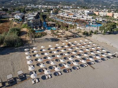 Playa con filas de sombrillas y tumbonas frente a un pueblo pequeño y colinas al fondo.