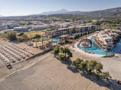 Hotel de lujo con gran piscina y tumbonas junto a playa de arena y montañas al fondo.
