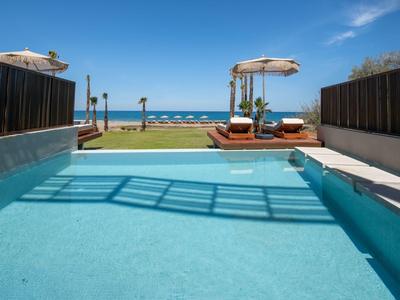 Čistý bazén s lehátky a slunečníky před mořem pod jasnou modrou oblohou.