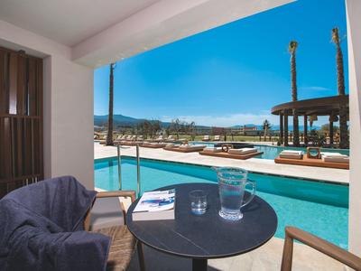 Balcony with table and chairs overlooking a pool and sun loungers under a blue sky.
