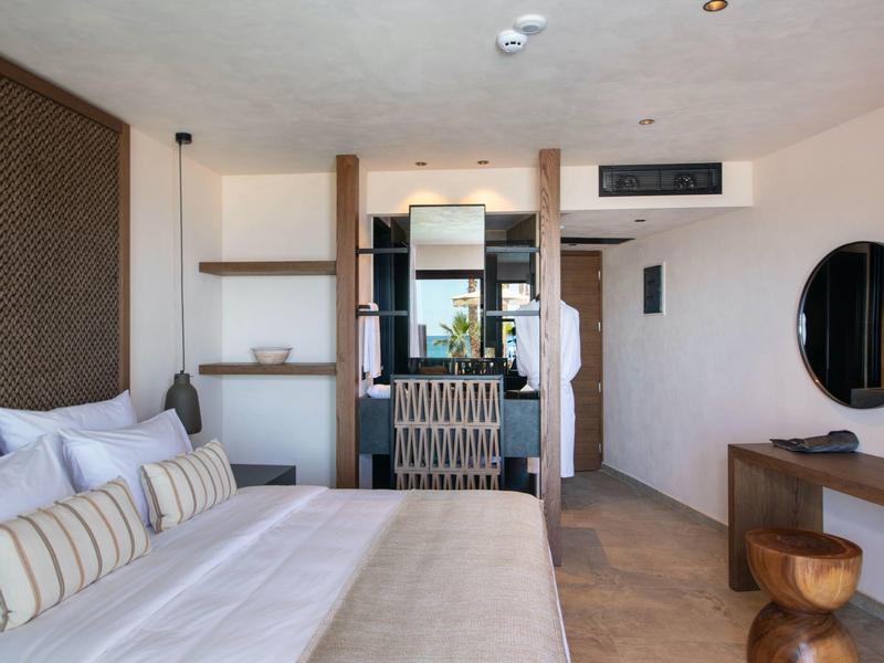 Moderní hotelový pokoj s velkou postelí, psacím stolem, kulatým zrcadlem a minimalistickým designem.