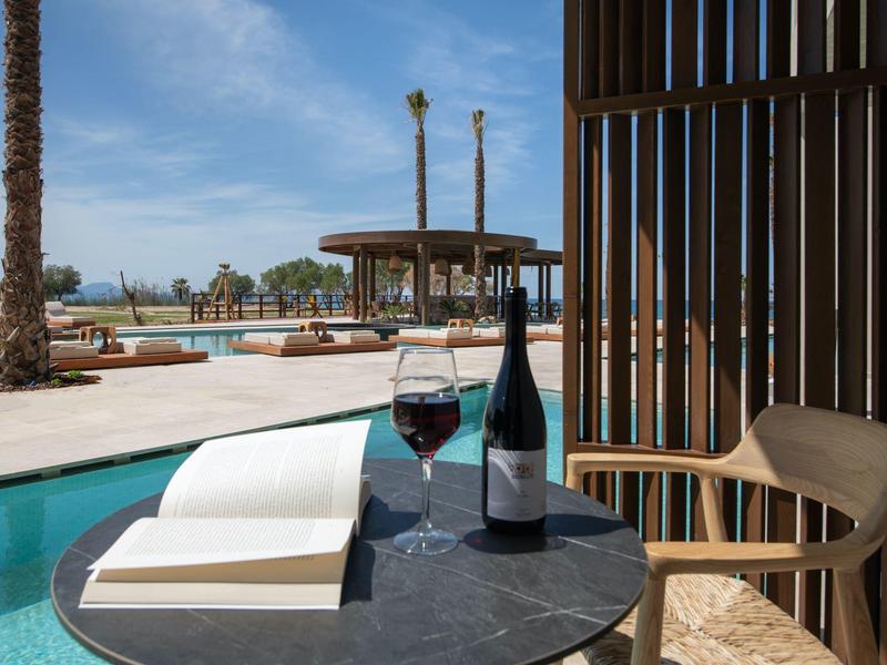 Terasa se židlí, kulatým stolem, lahví vína a sklenicí u bazénu pod modrou oblohou.