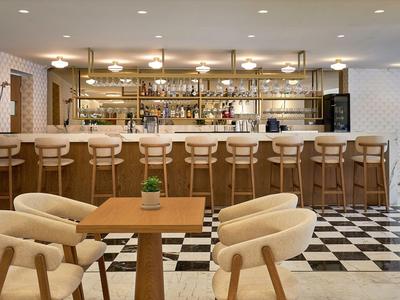 Moderner Hotelbarbereich mit hellem Holz, weißen Stühlen und schwarz-weißem Fliesenboden.