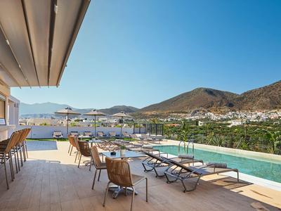 Terrasse mit Liegestühlen, Tischen und einem Pool mit Blick auf Hügel und Dorf unter klarem Himmel.