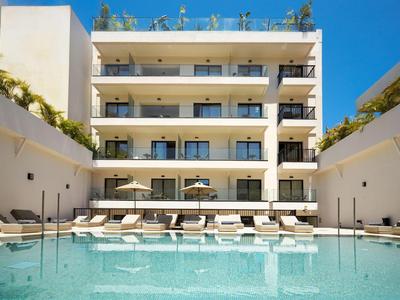 Moderner Hotelpool mit Liegestühlen vor einem mehrstöckigen Gebäude unter klarem Himmel.