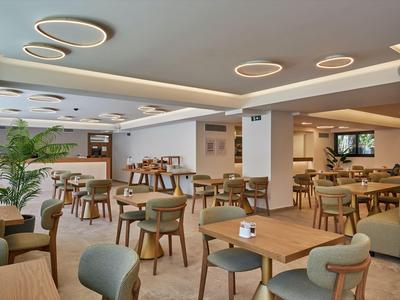 Moderner, heller Speisesaal mit Holztischen, gepolsterten Stühlen und Pflanzen in einem Hotel.