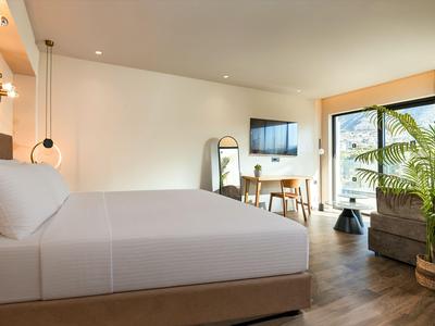 Modernes Hotelzimmer mit großem Bett, Schreibtisch, Fernseher und Balkon mit Pflanzen.