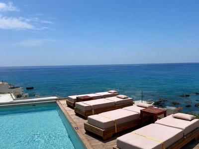 Pool mit Liegestühlen auf Holzdeck mit Blick auf das blaue Meer und klaren Himmel.