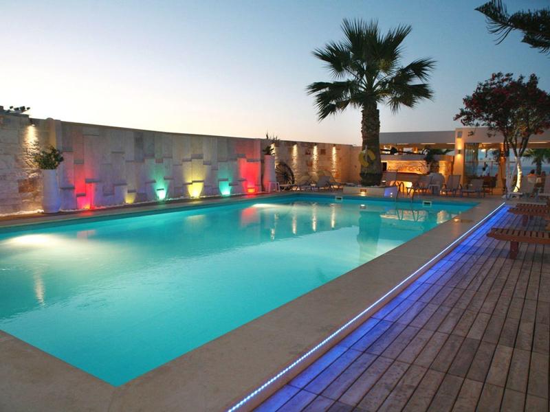 Beleuchteter Pool mit Holzterrasse, Palmen und farbigen Wandlichtern bei Dämmerung.