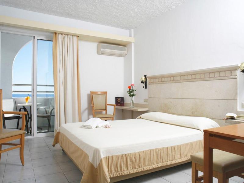 Helles Hotelzimmer mit Doppelbett, Holzmöbeln, Balkon und Blick aufs Meer.