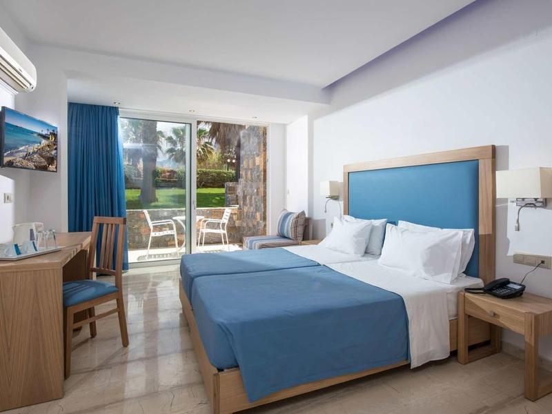 Světý hotelový pokoj s modrými přikrývkami a přístupem na terasu s výhledem do zahrady.