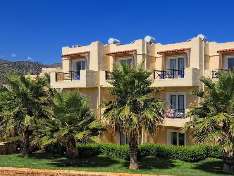 Hotelová budova s balkony a palmami před ní pod slunečnou oblohou.