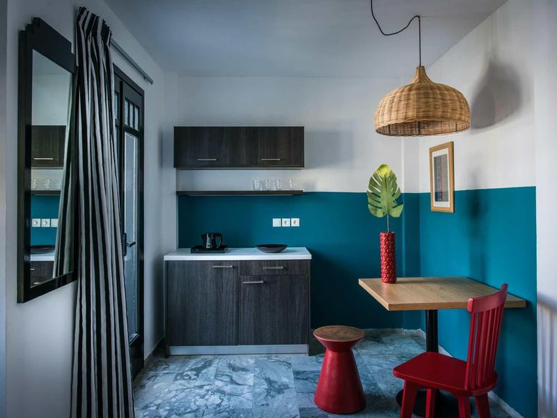 Piccola cucina con armadietti blu scuro, tavolo da pranzo, sedie colorate e decorazioni murali.