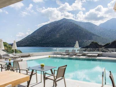 Area piscina moderna con sedie e tavoli davanti a una montagna e un lago sotto un cielo nuvoloso.