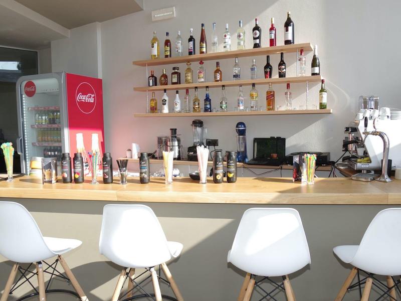 Bar mit weißem Tresen, vier weißen Stühlen, Regal mit Flaschen und einem roten Kühlschrank.