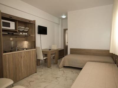 Kleines Zimmer mit zwei Betten, Holzküche, Tisch, Stuhl und Fenster mit hellem Vorhang.