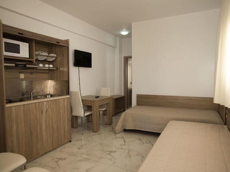 Kleines Zimmer mit zwei Betten, Holzküche, Tisch, Stuhl und Fenster mit hellem Vorhang.