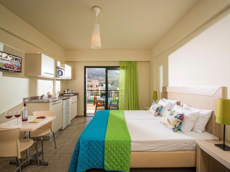 Modernes Hotelzimmer mit großem Bett, Küchenzeile, Esstisch, Balkon und grün-blauer Bettdecke.