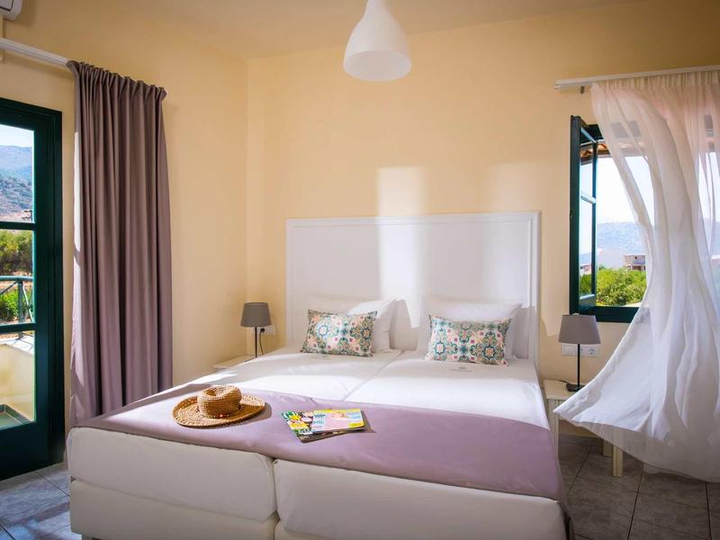 Helles Schlafzimmer mit Doppelbett, bunten Kissen, Fenster mit Vorhängen und Blick auf grüne Landschaft.