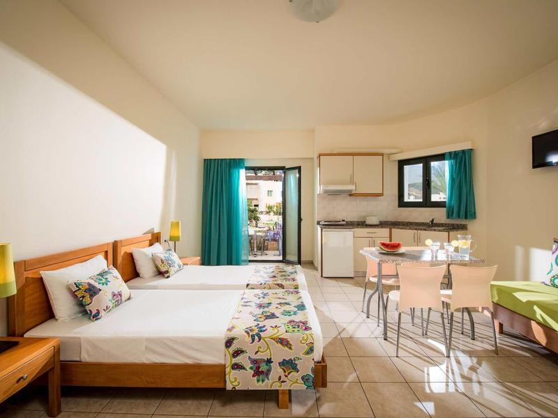 Hell eingerichtetes Zimmer mit Doppelbett, Küchenzeile, Esstisch, Sofa und Balkon.