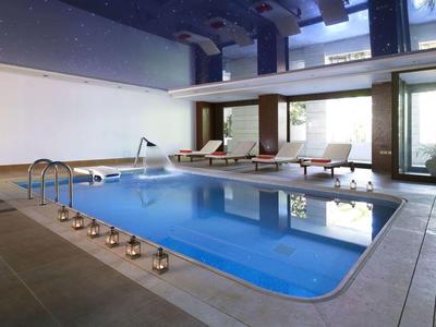 Moderner Innenpool mit Holzliegen, großen Fenstern und Kerzen am Beckenrand in hellem Raum.