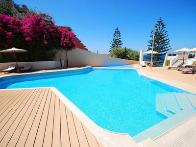 Area piscina limpida con lettini, ombrelloni e cespugli fioriti sotto un cielo azzurro.