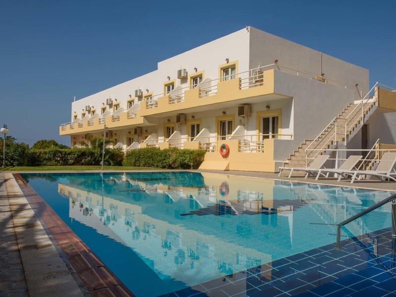 Moderner gelber Hotelbau mit Balkon und Klappstühlen neben großem blauem Schwimmbecken bei klarem Himmel.