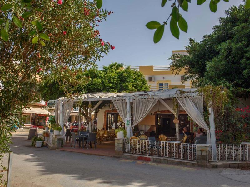 Offenes Restaurant mit hellen Vorhängen, Holzmöbeln und grünen Bäumen in sonniger Straße.