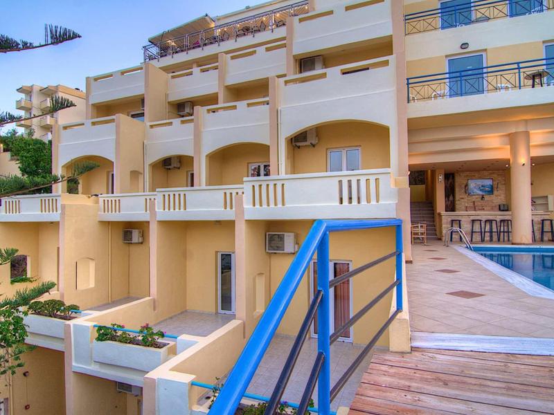 Mehrstöckiges gelbes Gebäude mit blauen Geländern und Pool auf einer Holzterrasse bei Sonnenuntergang.