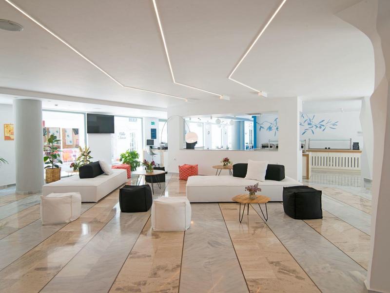 Hall d'hôtel moderne et lumineux avec canapés blancs, plantes et plafond géométrique.