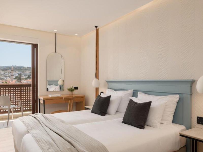 Bright hotel room with double bed, black pillows, and view of balcony and city.