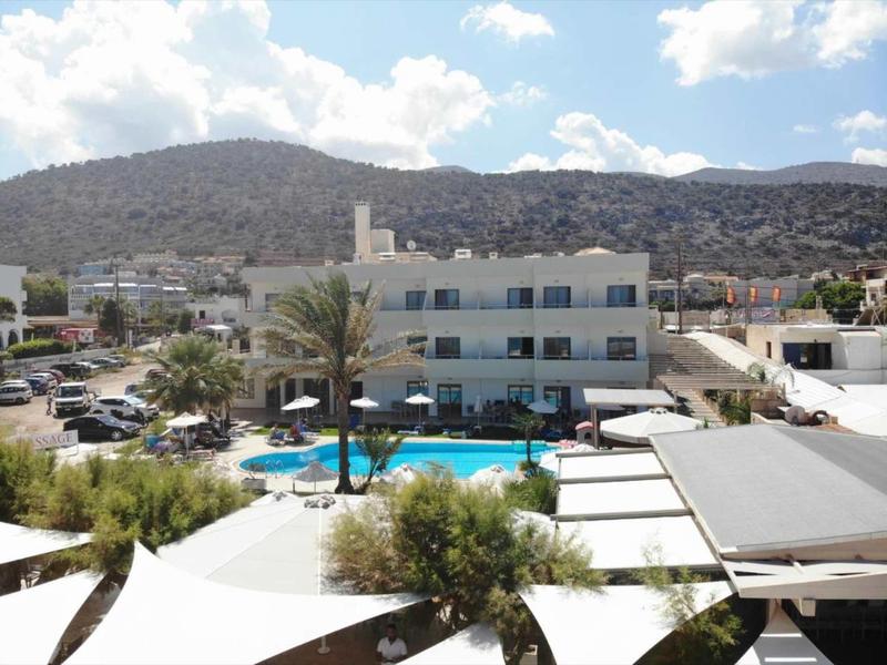 Hotel mit Pool, Palmen, mehreren Parkplätzen und bewaldeten Hügeln im Hintergrund unter blauem Himmel.