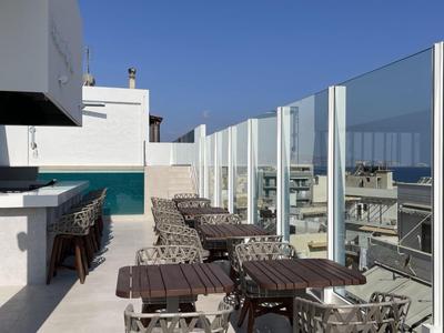 Moderní střešní terasa se stolky, židlemi a skleněnými zábradlími pod jasně modrou oblohou.