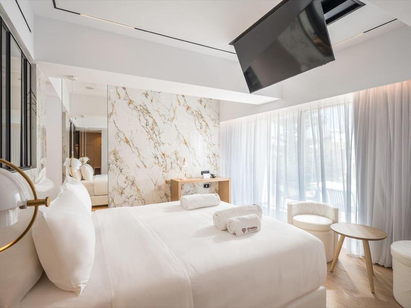 Camera d'albergo luminosa con letto bianco, parete in marmo e grande finestra con tende.