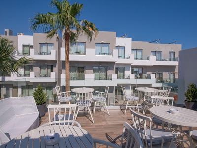 Terrasse mit weißen Tischen und Stühlen vor einem mehrstöckigen Hotelgebäude unter blauem Himmel.