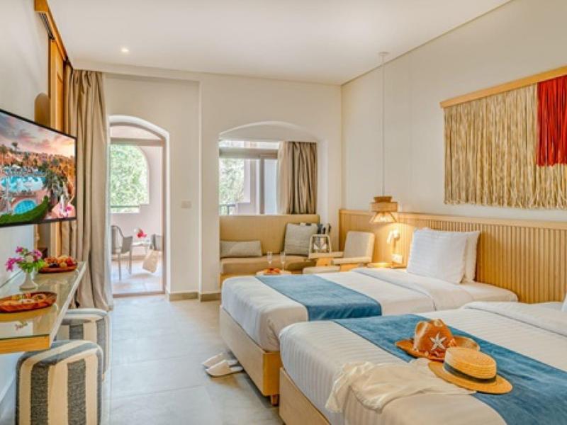 Lichte hotelkamer met twee bedden, bank, tv en balkon met zitje buiten.