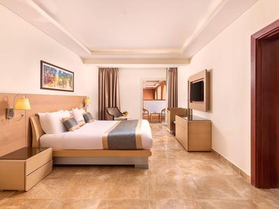 Habitación de hotel luminosa con cama grande, televisor, mesitas de noche y acceso a una segunda habitación con literas.