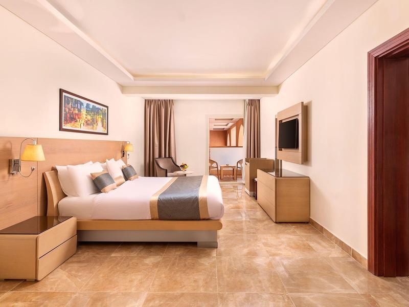 Habitación de hotel luminosa con cama grande, televisor, mesitas de noche y acceso a una segunda habitación con literas.