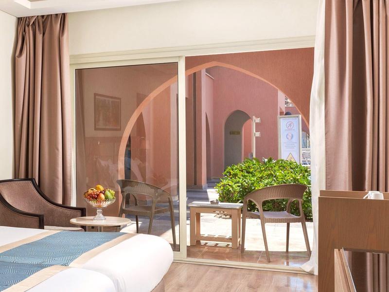 Habitación de hotel con vista a una terraza con plantas verdes y paredes rosas a través de una gran puerta corrediza de vidrio.