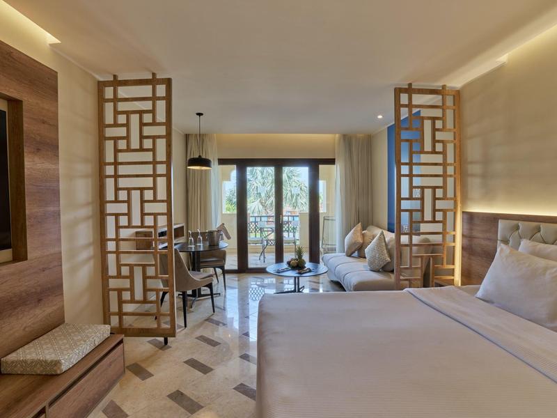 Chambre d'hôtel moderne avec grand lit, coin salon et portes donnant sur un balcon avec vue sur des plantes.