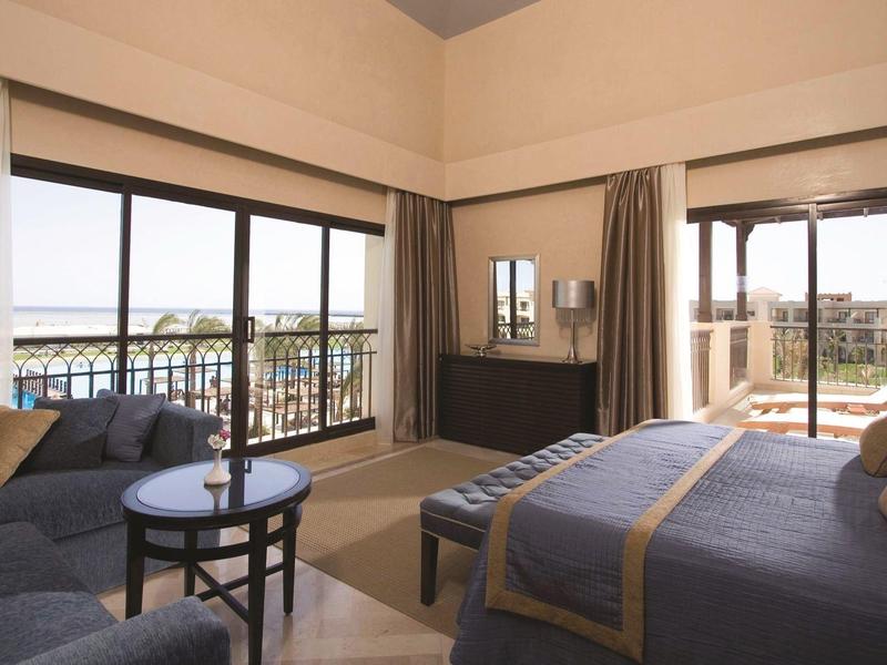 Hotelkamer met grote ramen, blauw beddengoed, banken en houten meubels met balkonzicht.